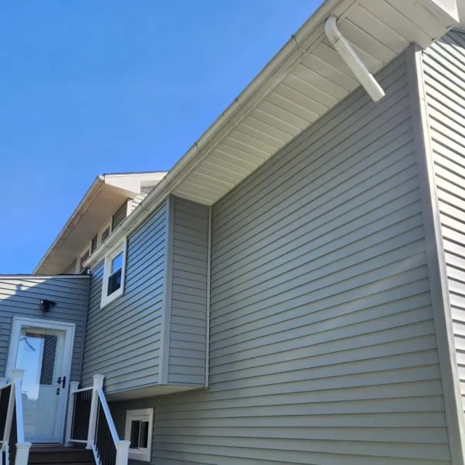 Side view of a house with gray siding, white gutters, and a blue sky. A door and stairs are visible.