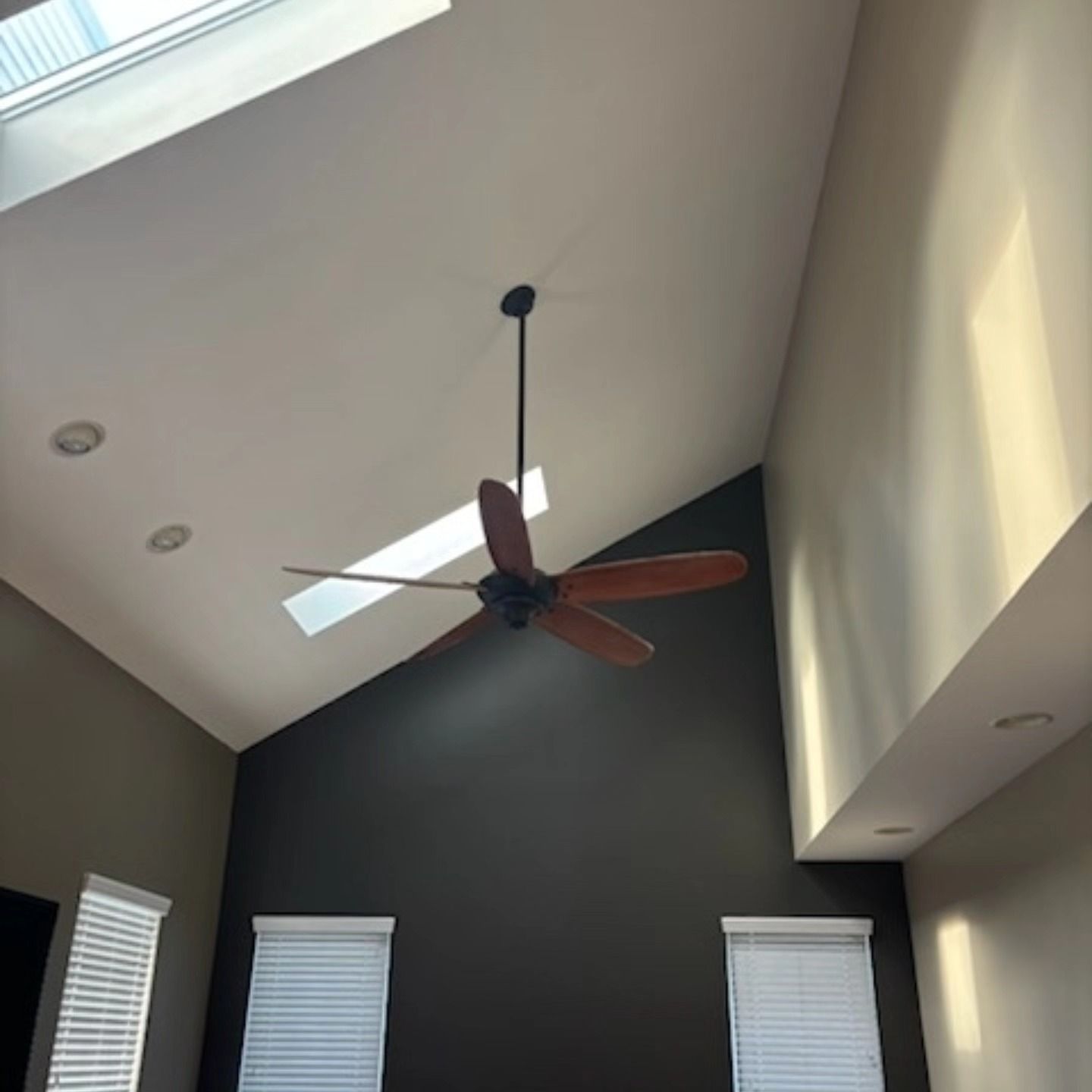 High-ceilinged room with a ceiling fan. Skylights and windows. Dark accent wall, beige and white walls.