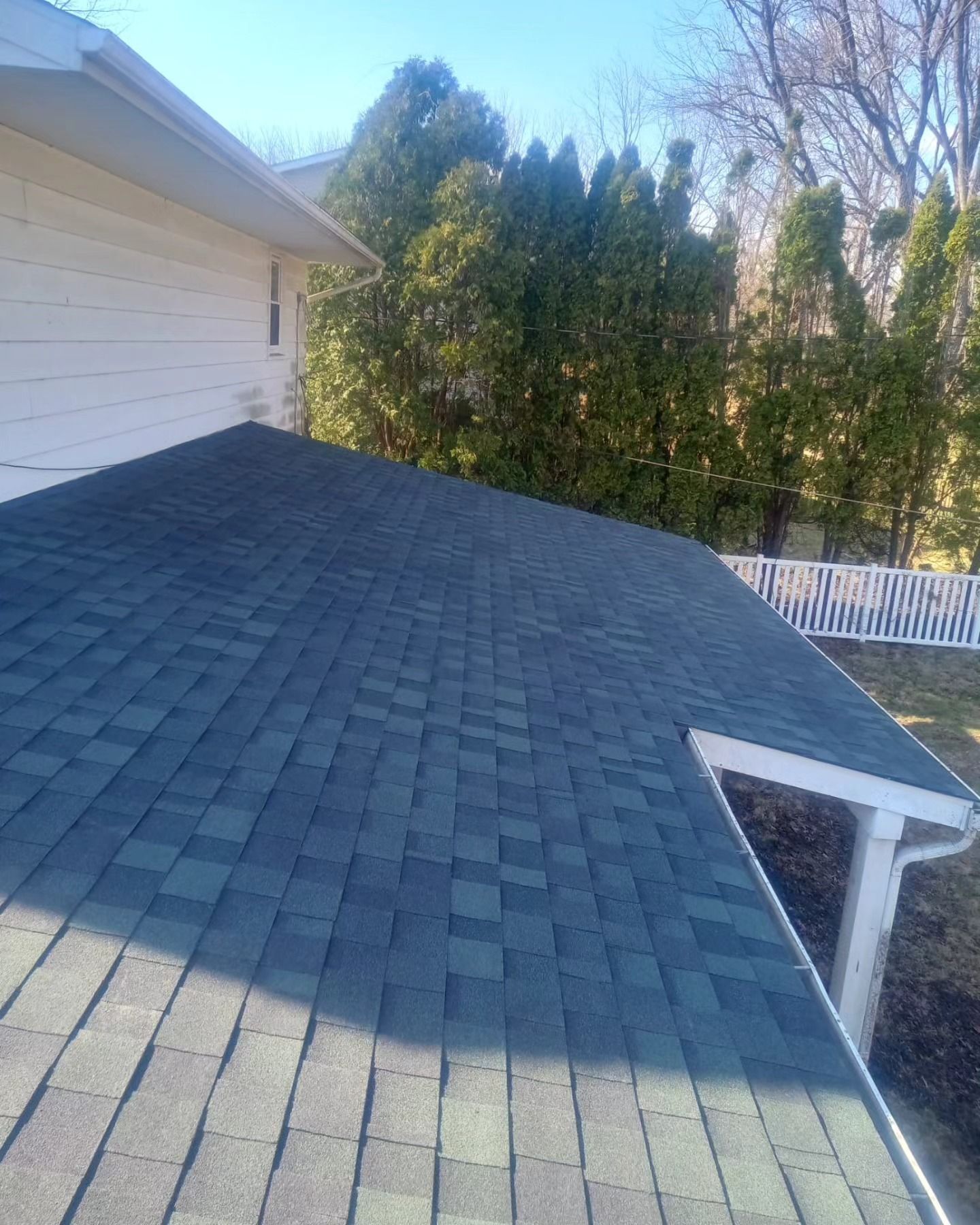 A dark gray shingle roof on a white house, with trees and a white picket fence in the background.