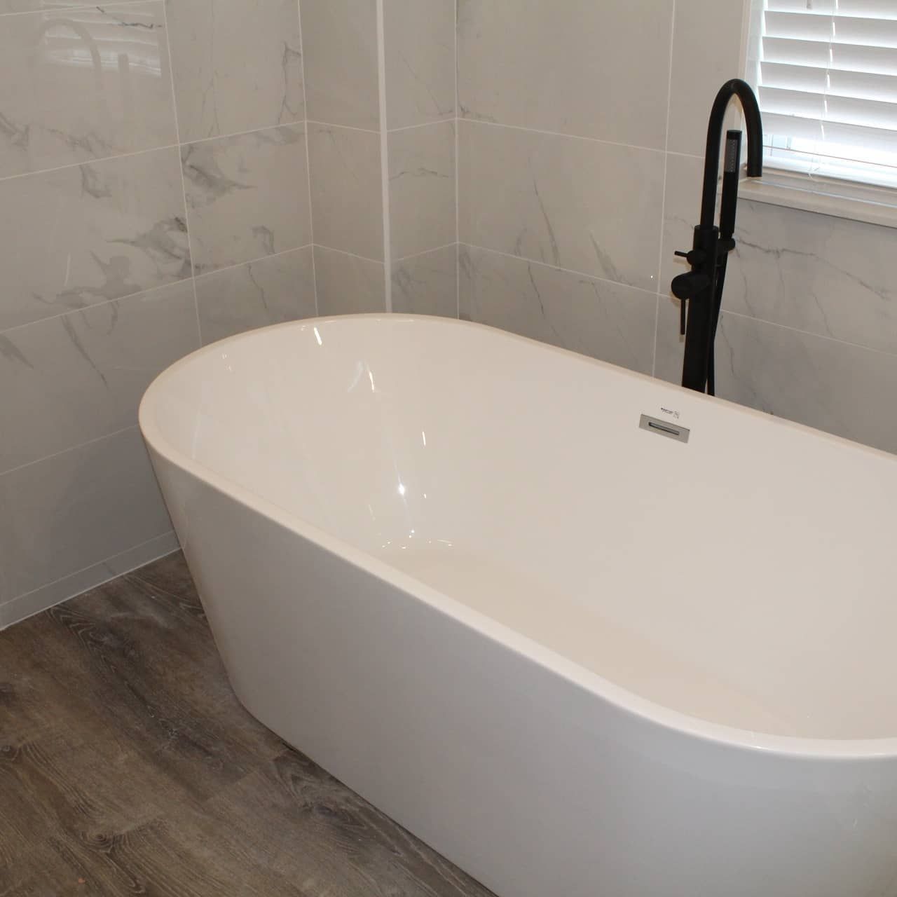 White soaking tub in a bathroom with gray marbled tile walls, a black faucet, and wood-look flooring.