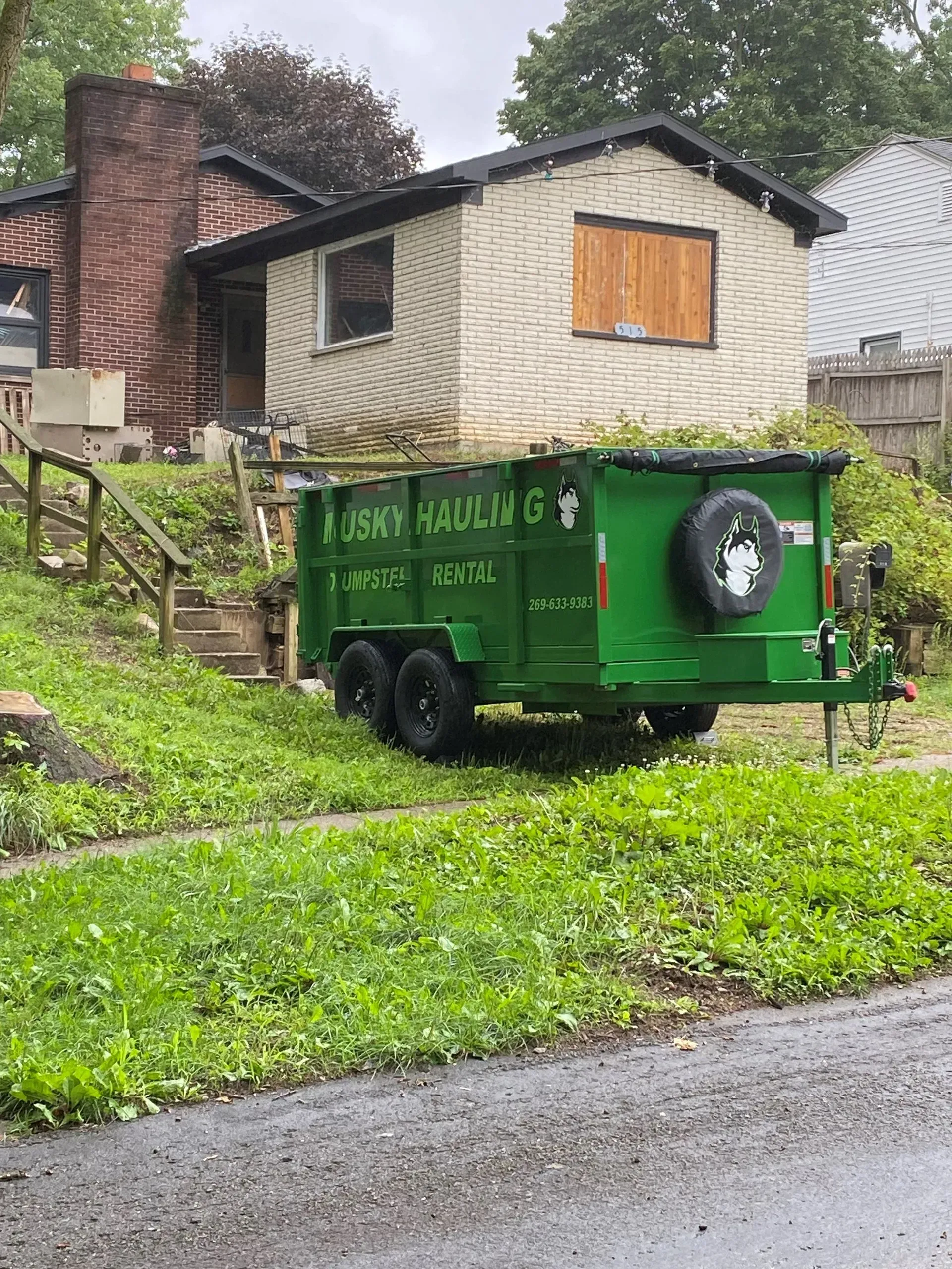 Green dumpster trailer parked on a residential lawn in front of a house.