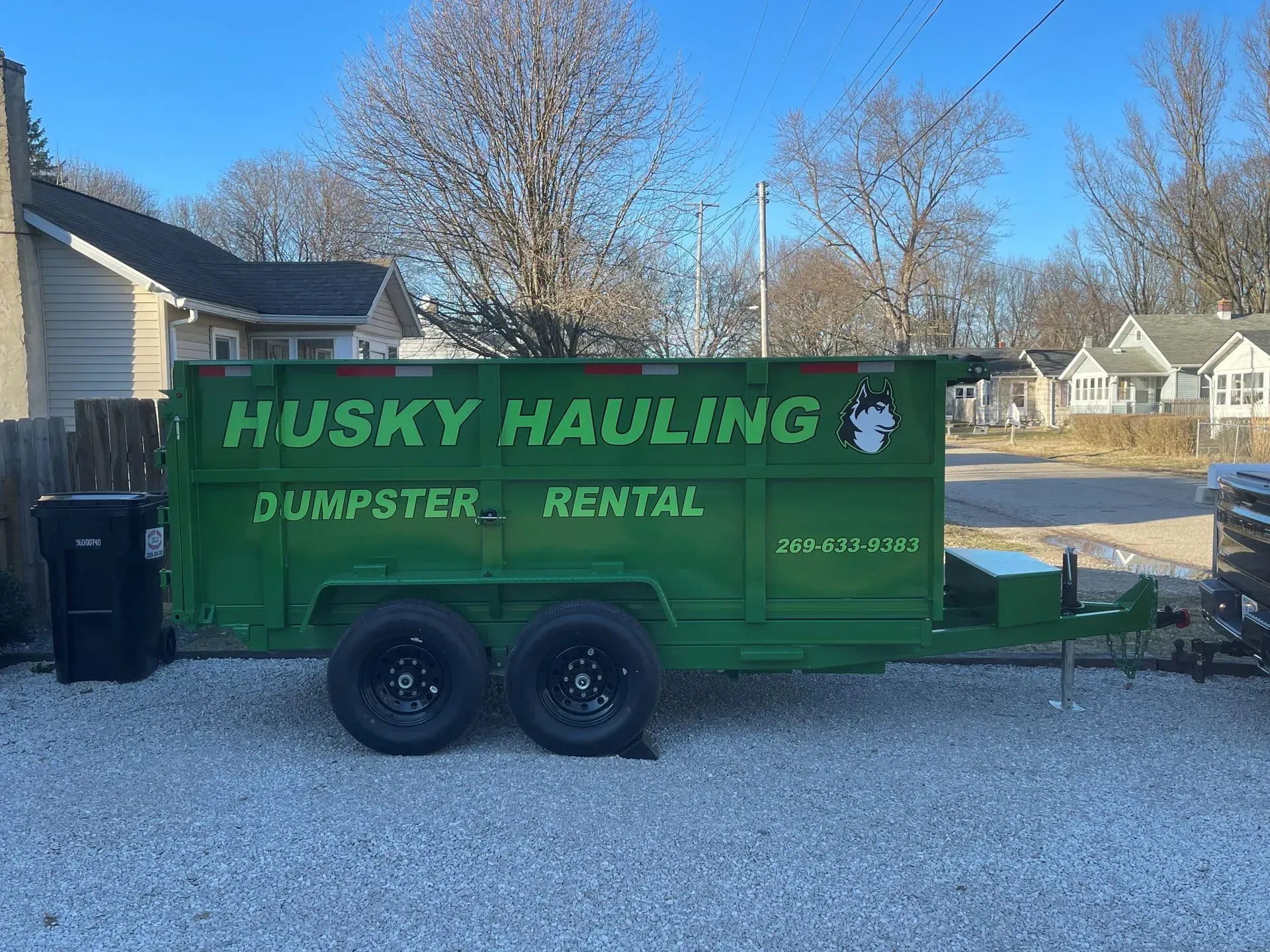 Green Husky Hauling dumpster trailer on gravel, parked near buildings.