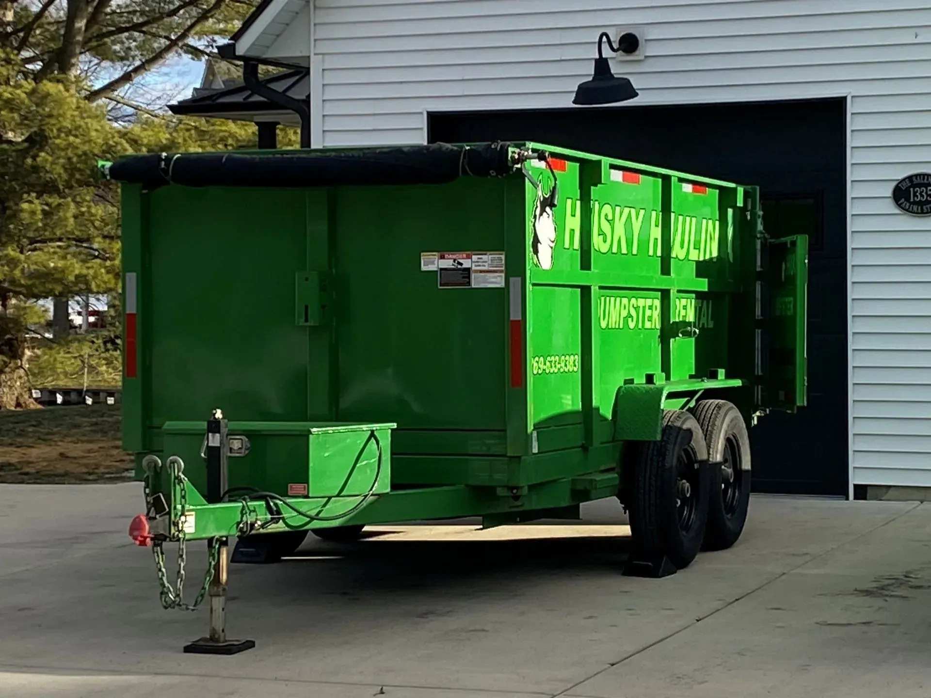 Green dumpster trailer parked in front of a white garage. The trailer reads