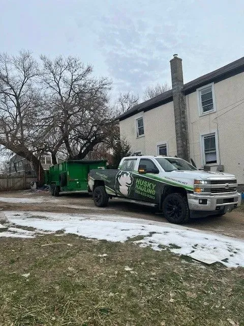 White and green truck with trailer in front of a beige building, tree, and snow-covered ground.