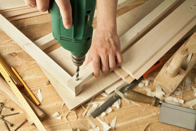 Person drilling a wooden board on a workbench with scattered tools and wood shavings.