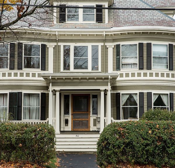 Victorian-style house facade with white trim, large windows, and a front porch behind hedges.