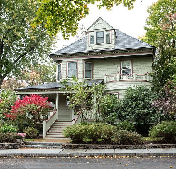 Tree-lined view of a pale green Victorian house with front porch, dormer, and landscaped yard.