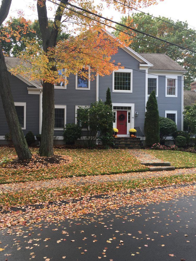 Gray house with white trim and red door behind autumn trees and fallen leaves on a quiet street.