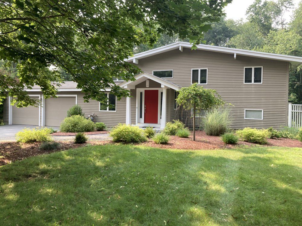 Suburban house with tan siding, red front door, white garage, and landscaped yard under trees.
