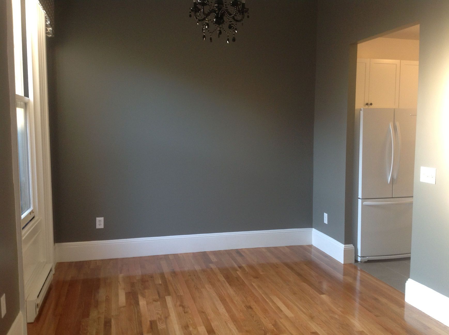 Empty dining room with hardwood floors, gray walls, white trim, and a refrigerator visible through the doorway.