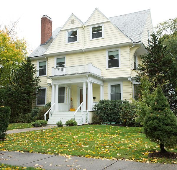 White two-story house with porch, red brick chimney, and a green lawn with fallen leaves.