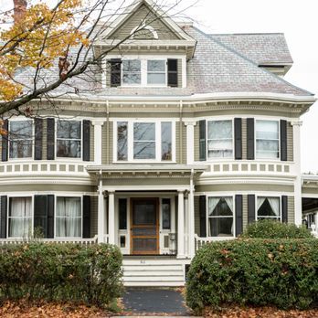 Victorian-style house with white trim, gray siding, and a front porch behind hedges and autumn tree branches.