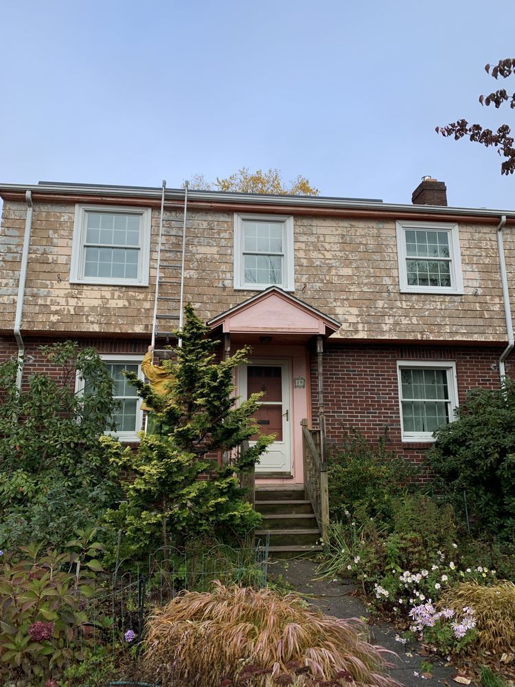 Two-story brick house with a pink awning over the front door, surrounded by shrubs and flowers.