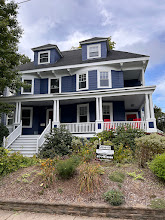 Blue two-story house with white trim, front porch, stairs, and landscaping under a cloudy sky.