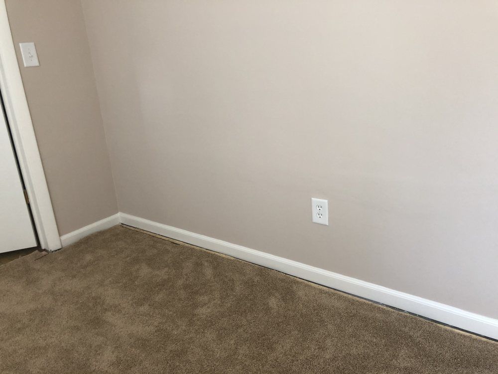 Empty beige room corner with brown carpet, white baseboards, and a door on the left.