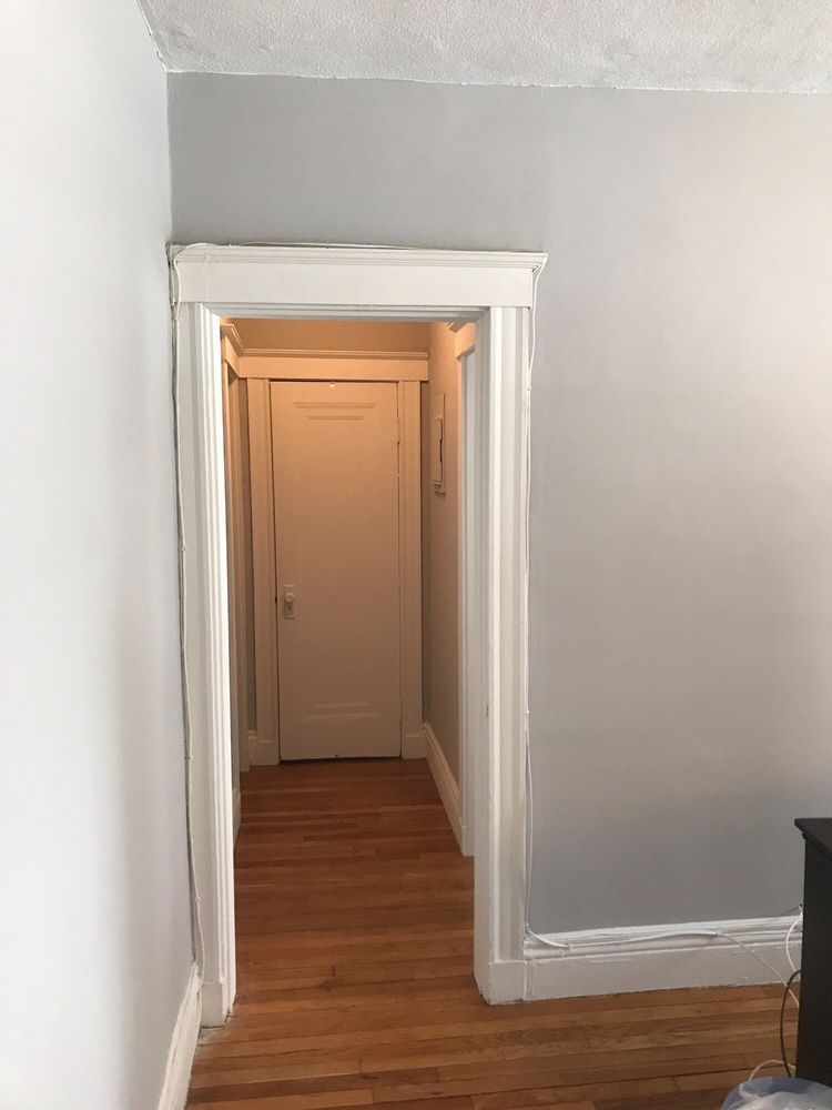 White hallway with wood floor leading to a closed beige door through a narrow doorway.