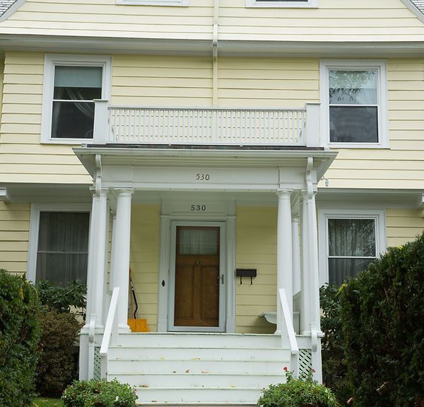 Front porch of a pale yellow house with white columns, wooden door, and balcony above the entrance.