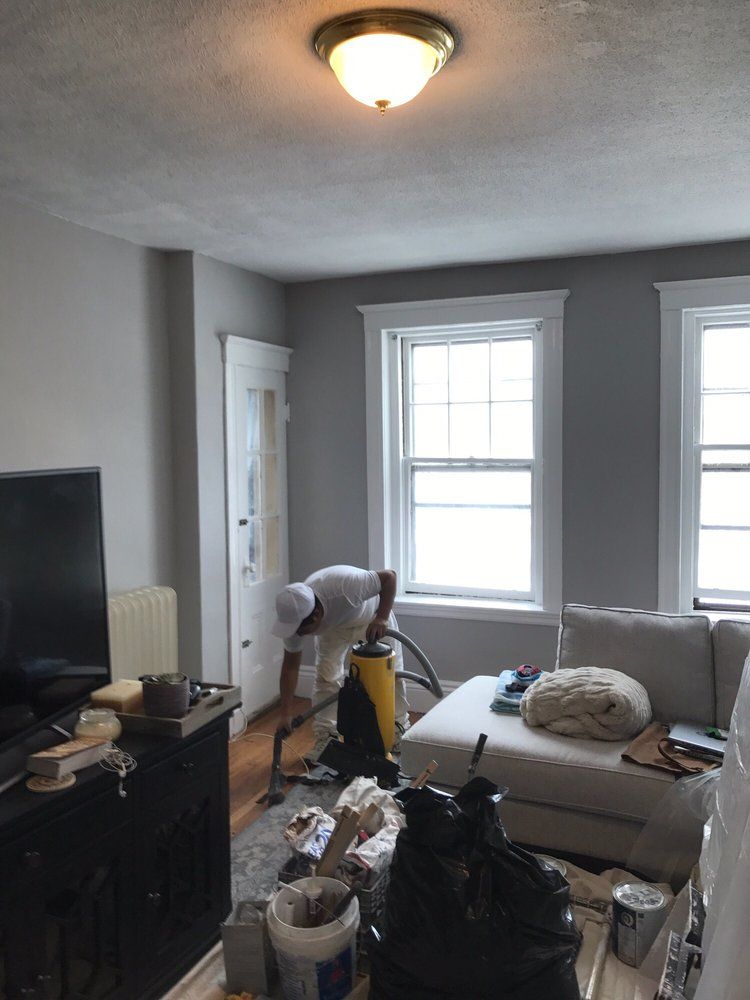 Cluttered gray bedroom with twin windows, a bed, TV, and cleaning supplies piled in the foreground.