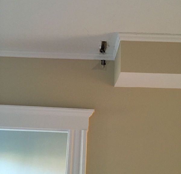 Close-up of a room corner with beige walls, white trim, and a small bracket mounted near the ceiling.