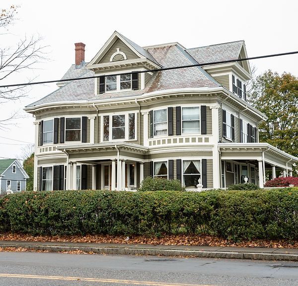 Large Victorian-style house with gray roof, white trim, and wraparound porch behind a hedge.