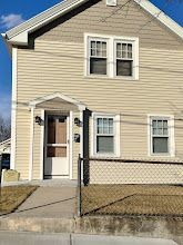 Beige two-story house with white trim, small front porch, and chain-link fence along the sidewalk.