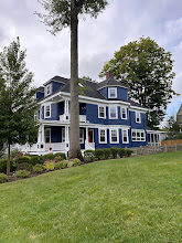 Blue colonial house with white trim on a grassy hill, framed by tall trees and a cloudy sky