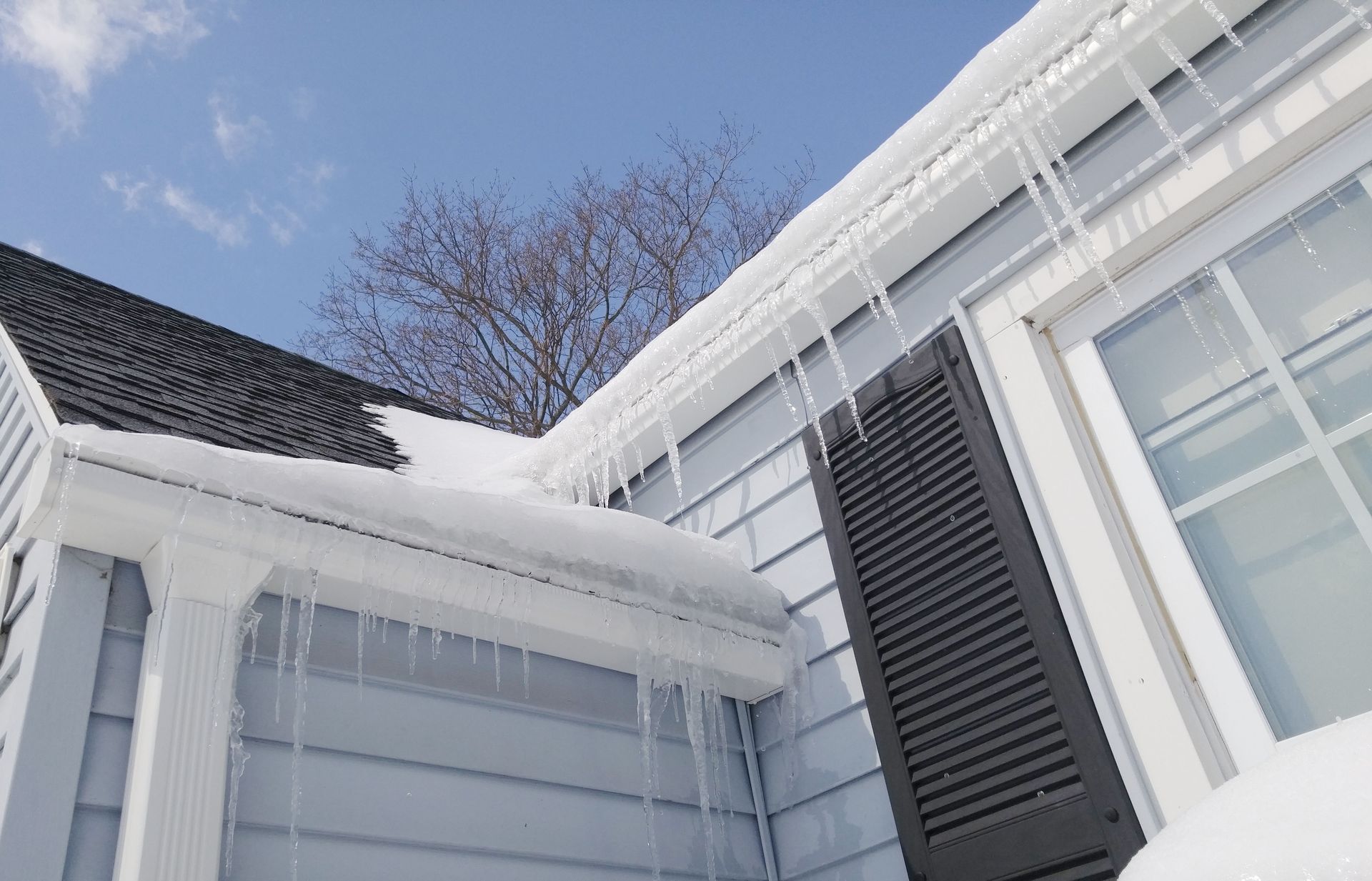Snow-covered house exterior with white siding, gray shutters, and bare trees against a blue sky
