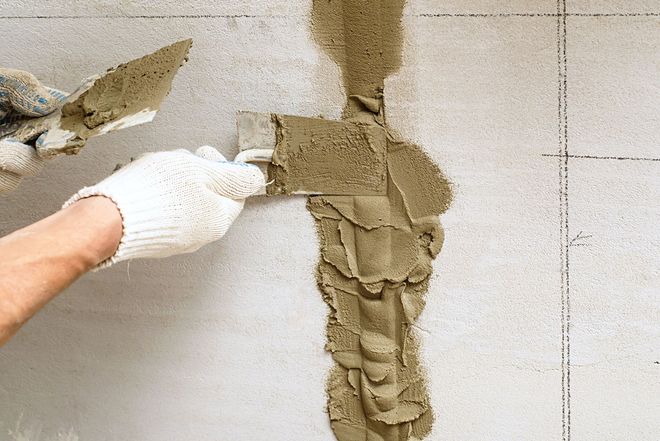 Gloved hand spreading wet plaster on a tiled wall repair.