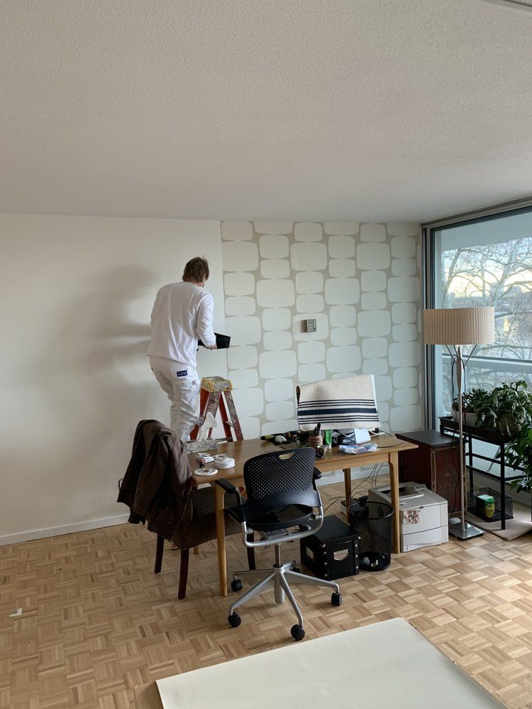 Person painting a white wall in a bright room with a desk, chair, ladder, and large window.