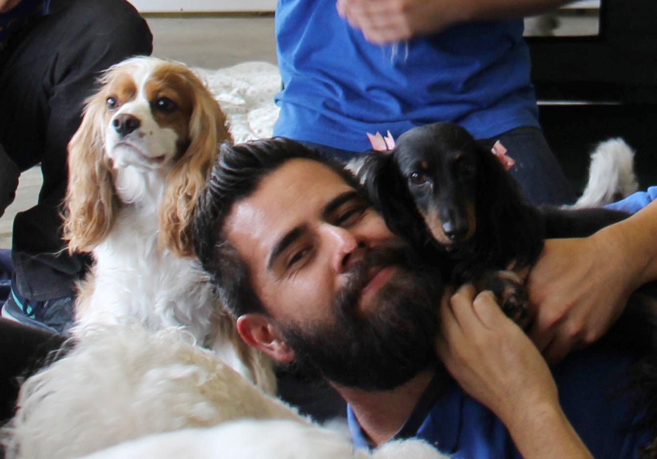 A man with a beard holds a small black dog while a Cavalier King Charles Spaniel sits beside him.