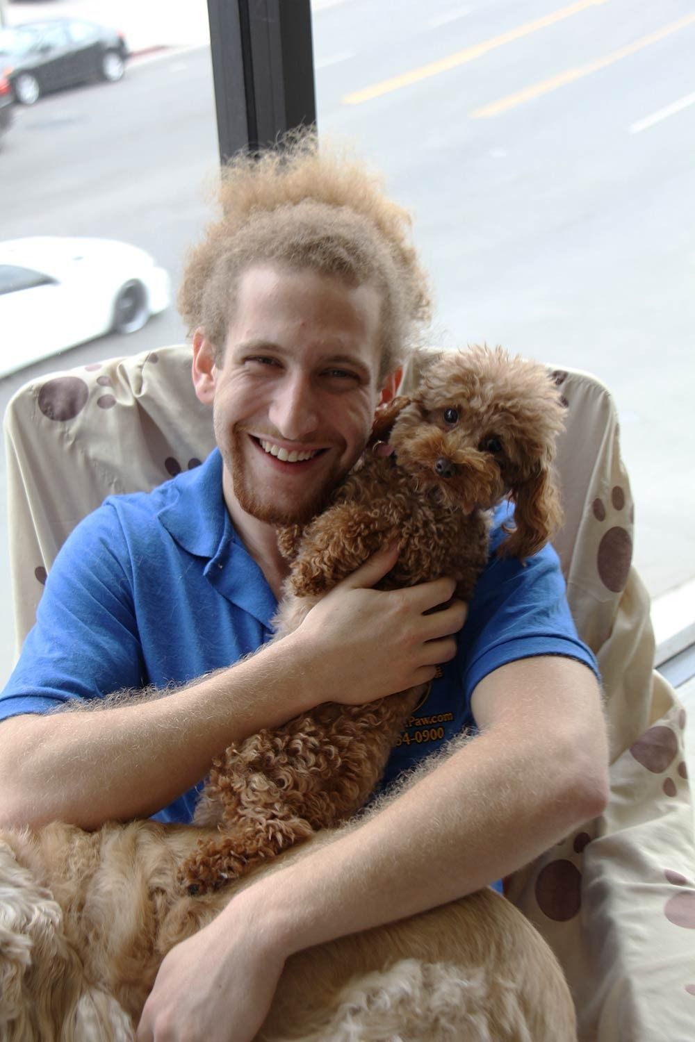 A person with curly hair smiling while holding a small, brown, curly-haired dog in a chair with a paw-print blanket.