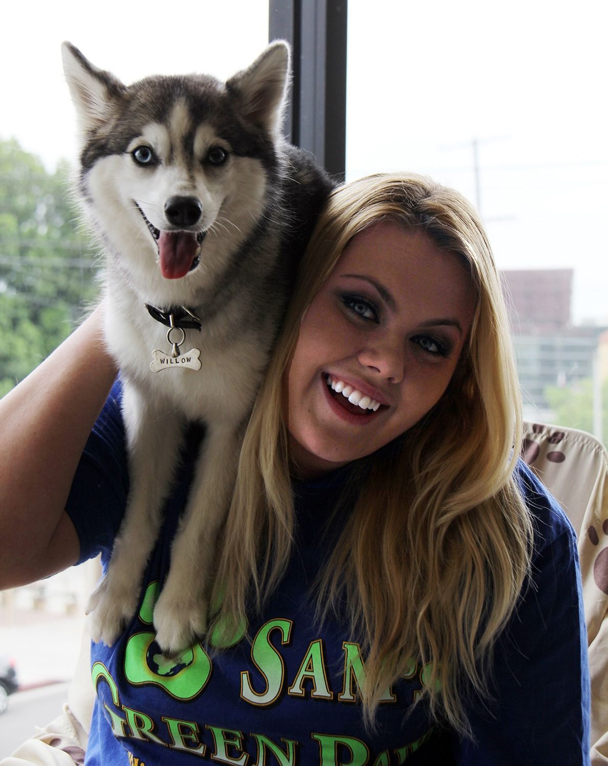 A smiling woman with long blonde hair holds a happy Siberian Husky on her shoulder in front of a window.