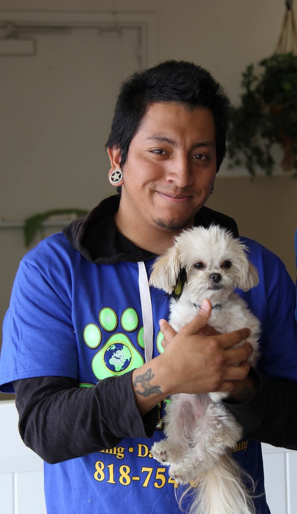A person in a blue shirt with a dog-themed logo holding a small, fluffy white dog.