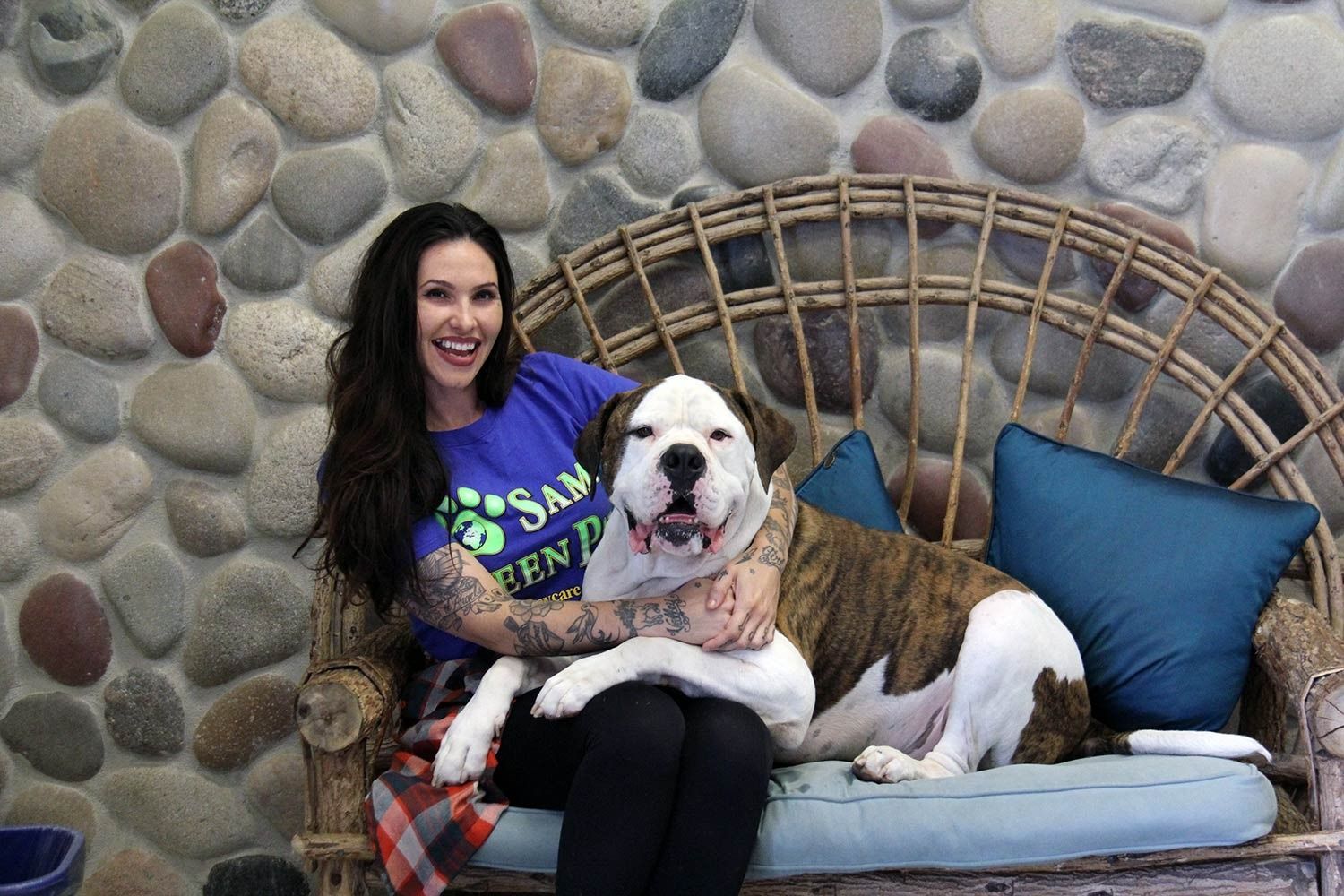 A person with arm tattoos wearing a blue t-shirt sits on a rattan sofa next to a brindle and white dog against a stone wall.