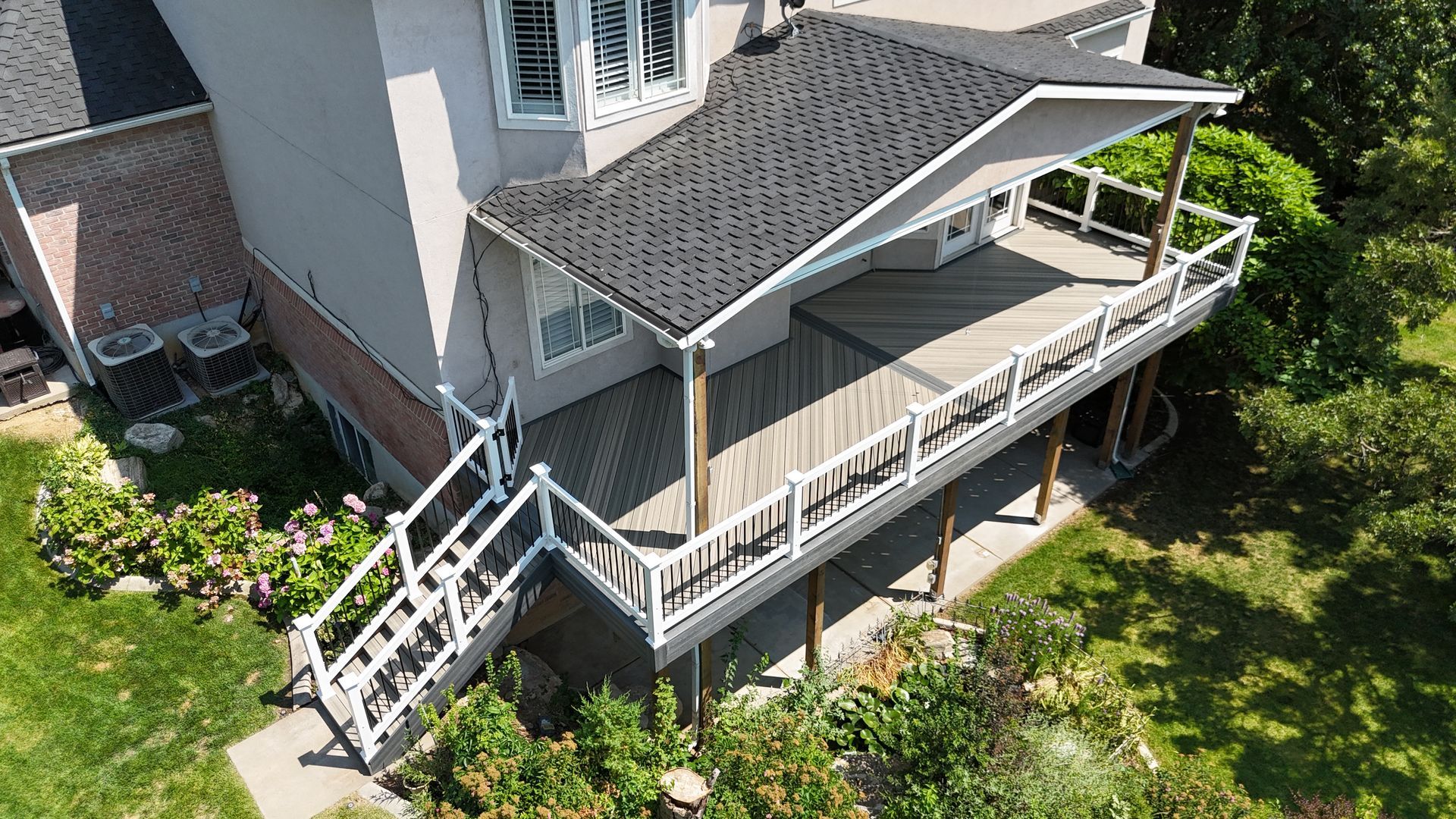 A house with a wraparound wooden deck, white railings, and stairs beside a green garden.