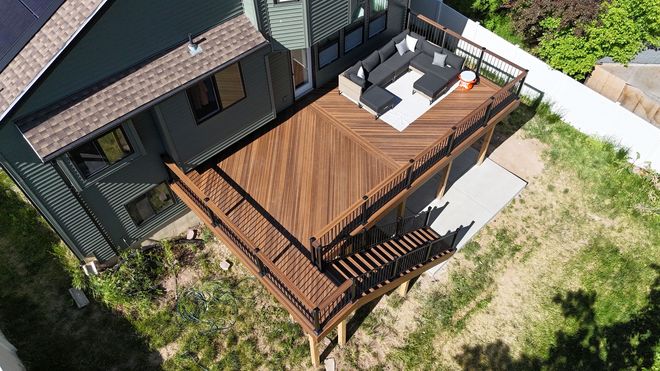 Multi-level light wood deck attached to a gray house, surrounded by lush green trees in a sunny backyard.