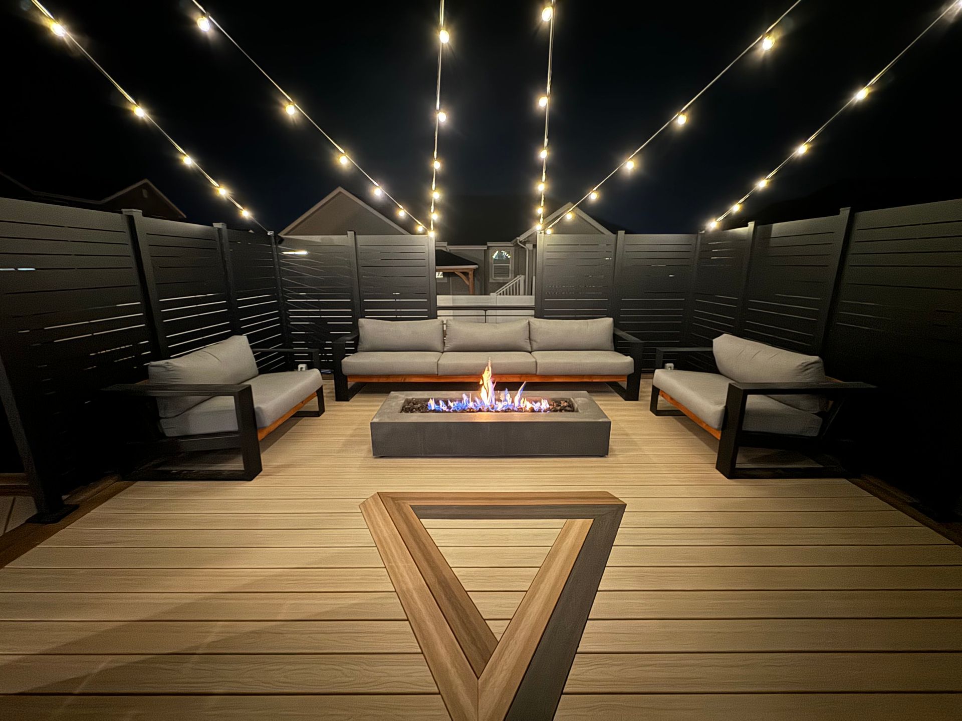Outdoor deck lounge with string lights, cushioned seating, and a central fire pit at night
