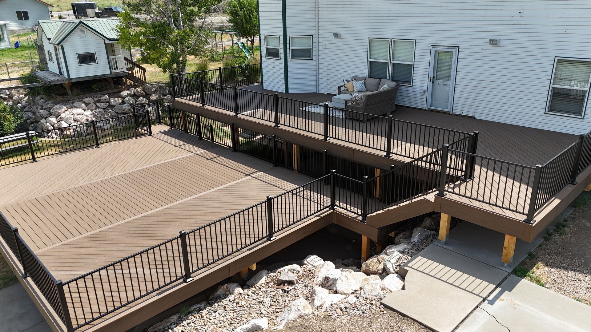 Brown wooden deck with railings beside a white house, featuring a raised platform and stairs.