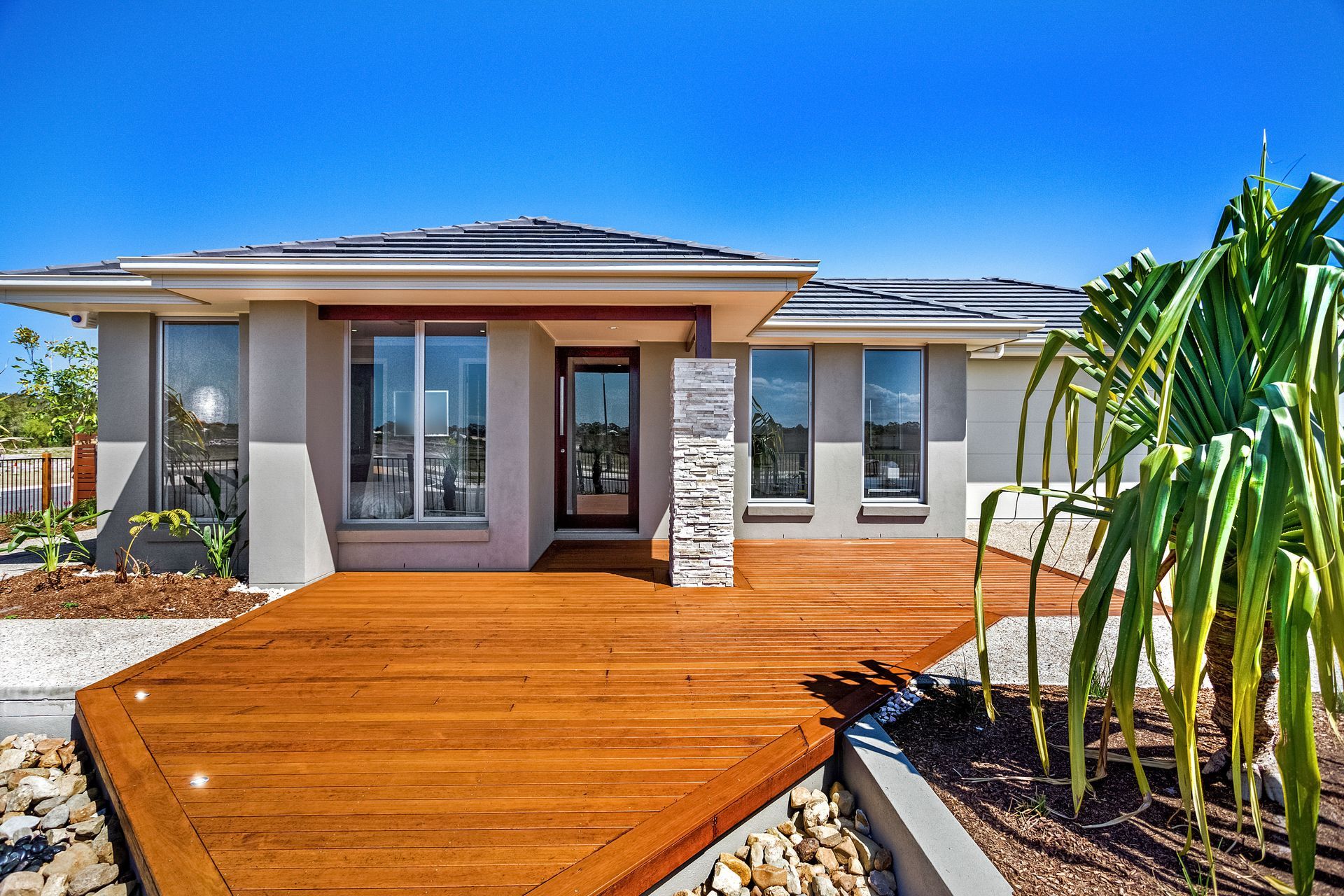 Modern single-story house with a wooden front deck and tropical landscaping under a blue sky