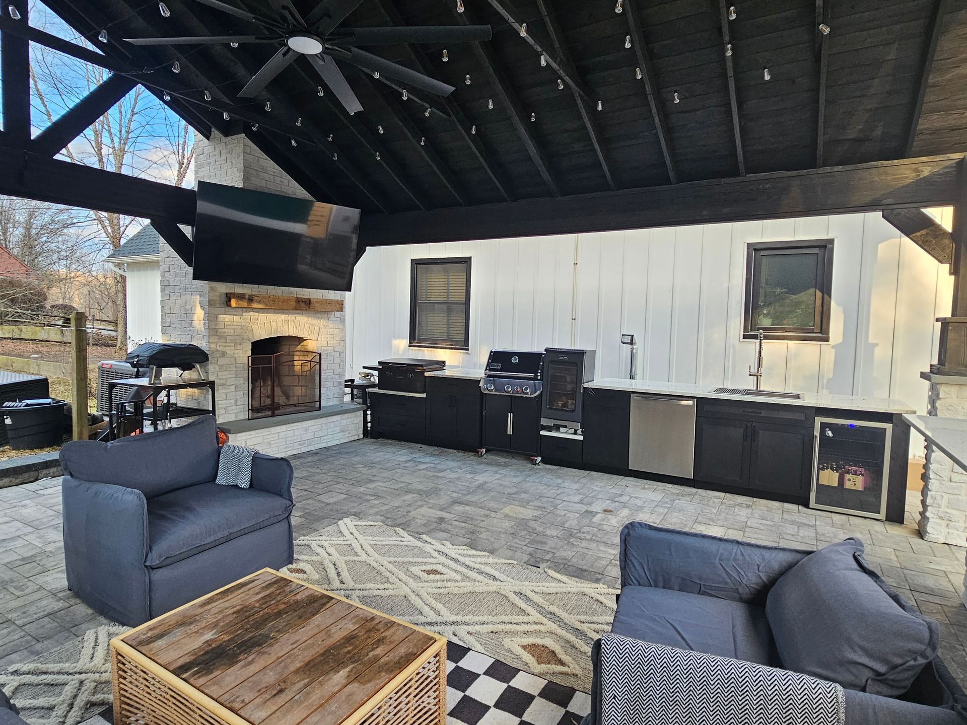 Covered patio with gray chairs, wood coffee table, grill, fireplace, and outdoor kitchen under a dark vaulted roof