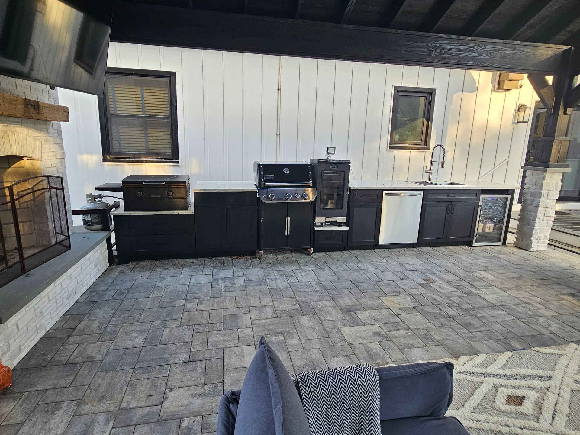 Outdoor patio kitchen with black cabinets, grill, and stone flooring under a covered roof