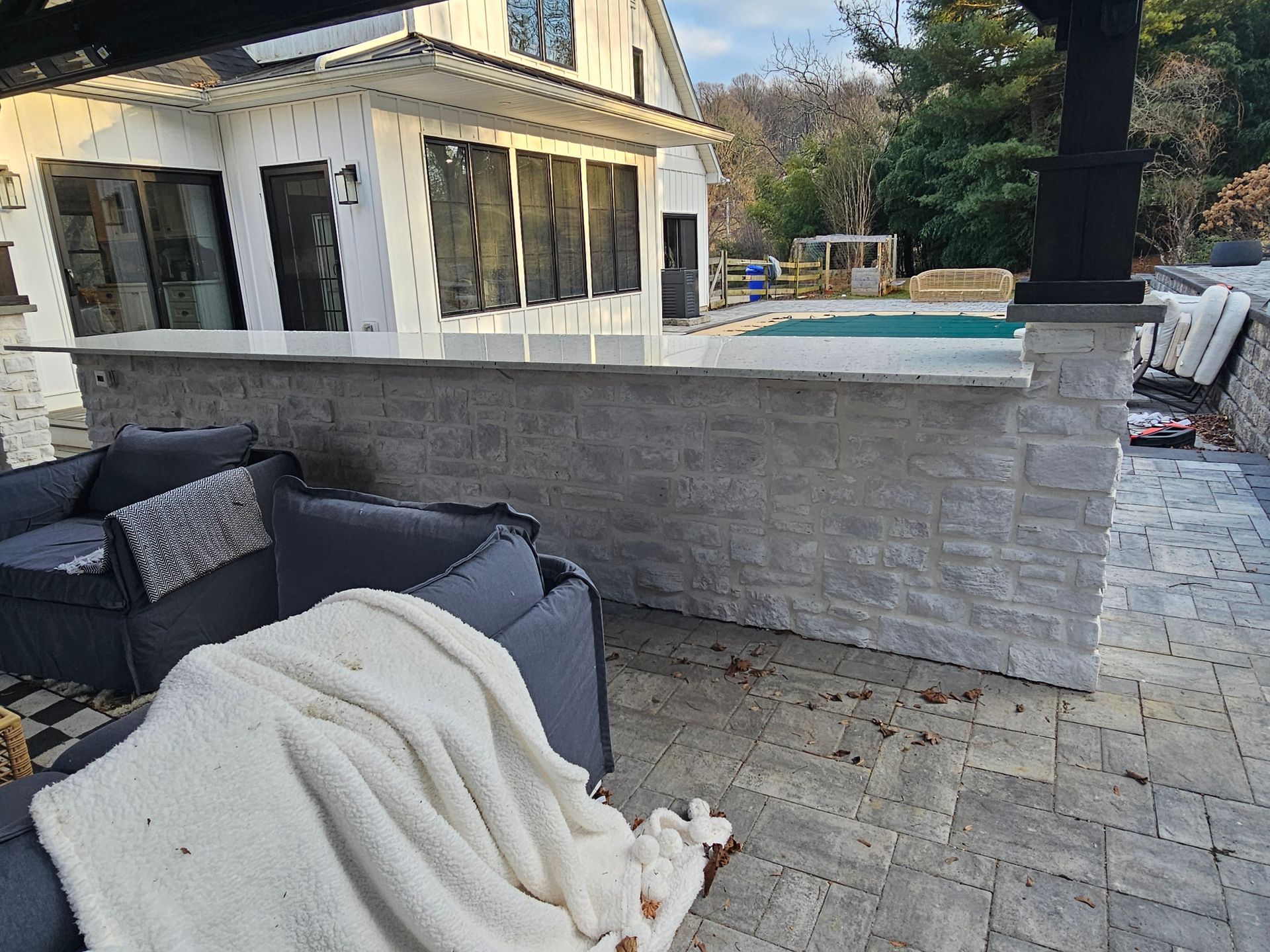 Patio with gray outdoor sofa and blanket beside a house and pool, seen in daylight