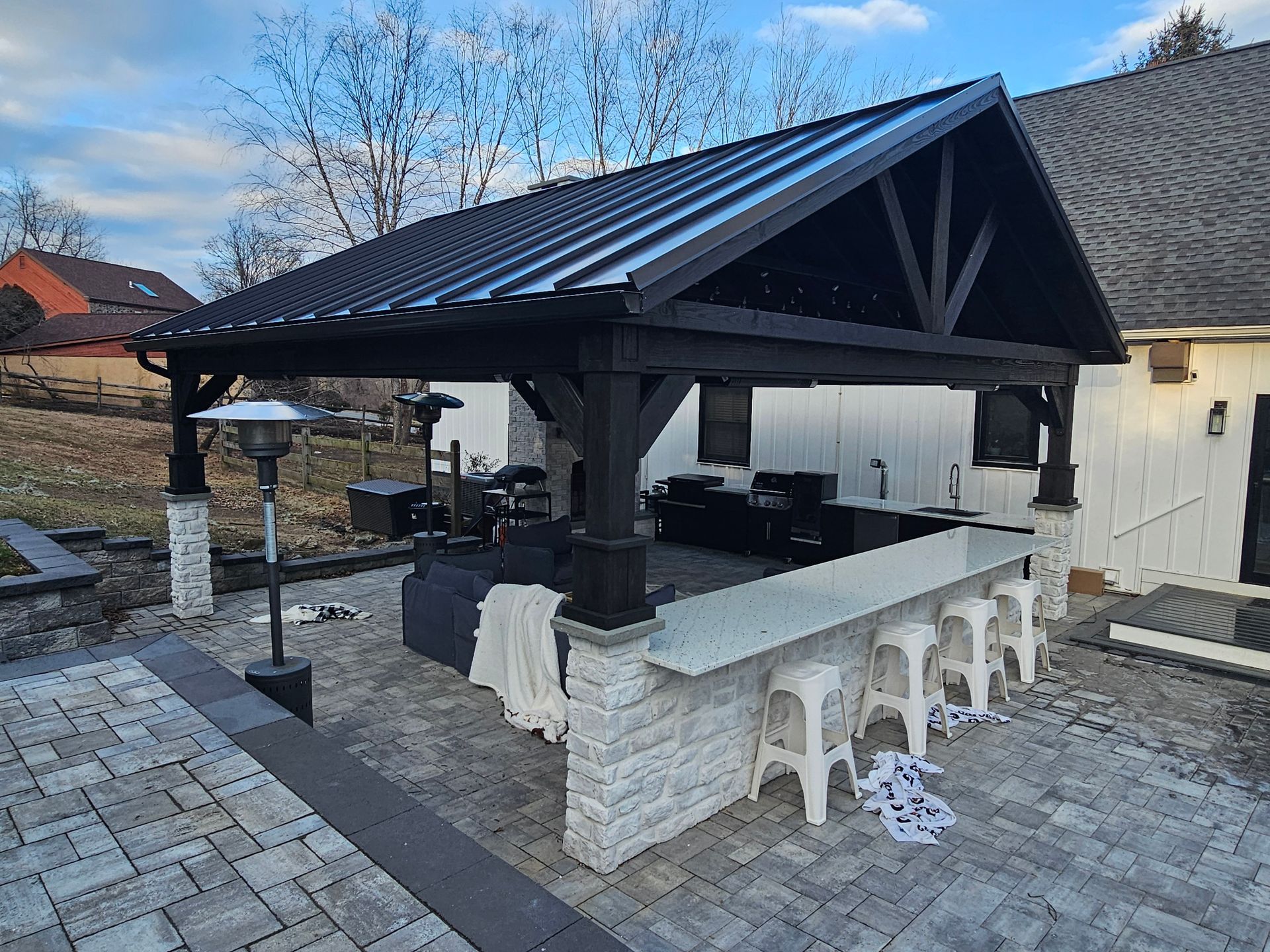 Covered outdoor bar with stone counter and stools under a dark wooden pavilion in a yard.