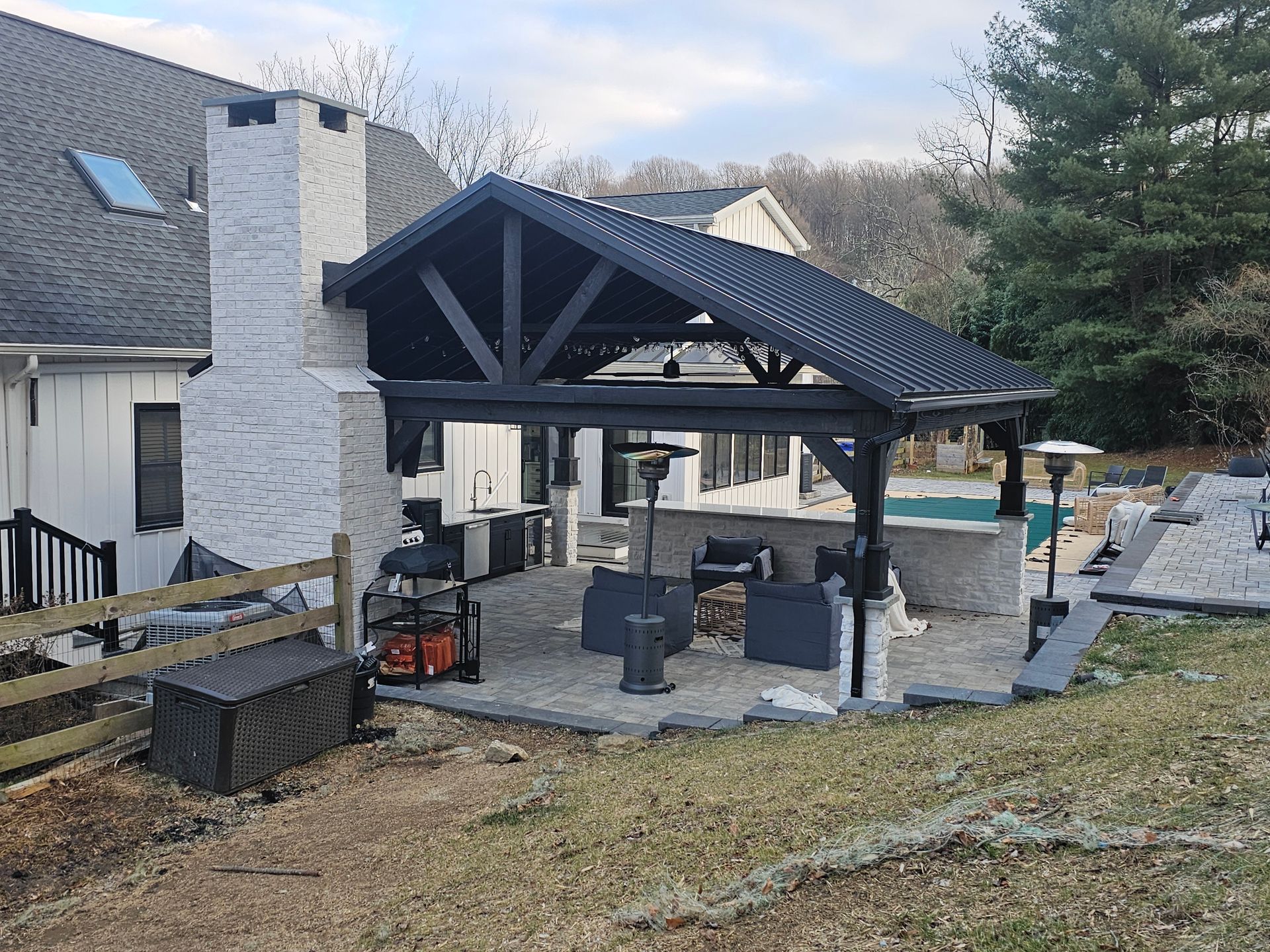Covered patio with dark roof, stone fireplace, and outdoor seating beside a house and pool.