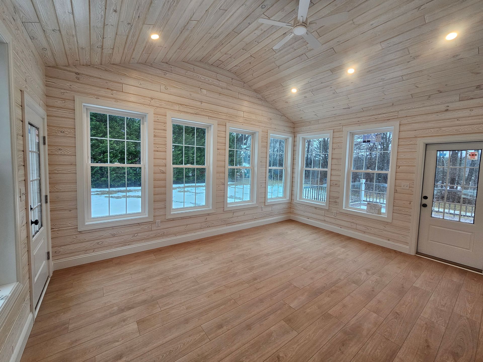 Empty sunroom with wood floors, white paneled walls, and large windows overlooking snowy trees