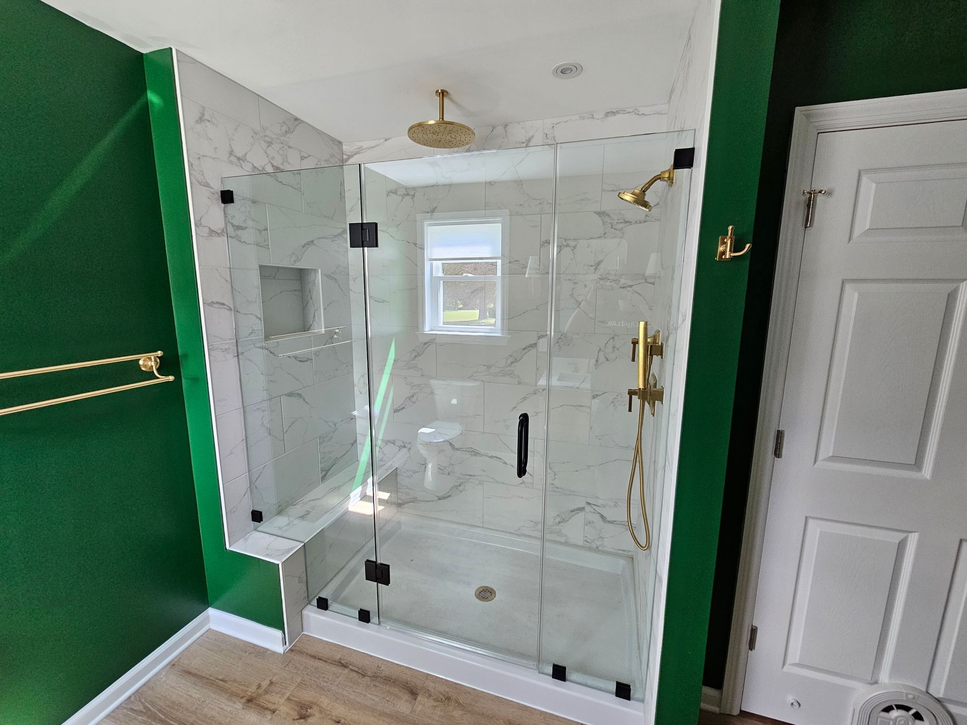 Newly renovated bathroom with a glass shower, marble tile, and gold fixtures. Green walls and a white door are visible.