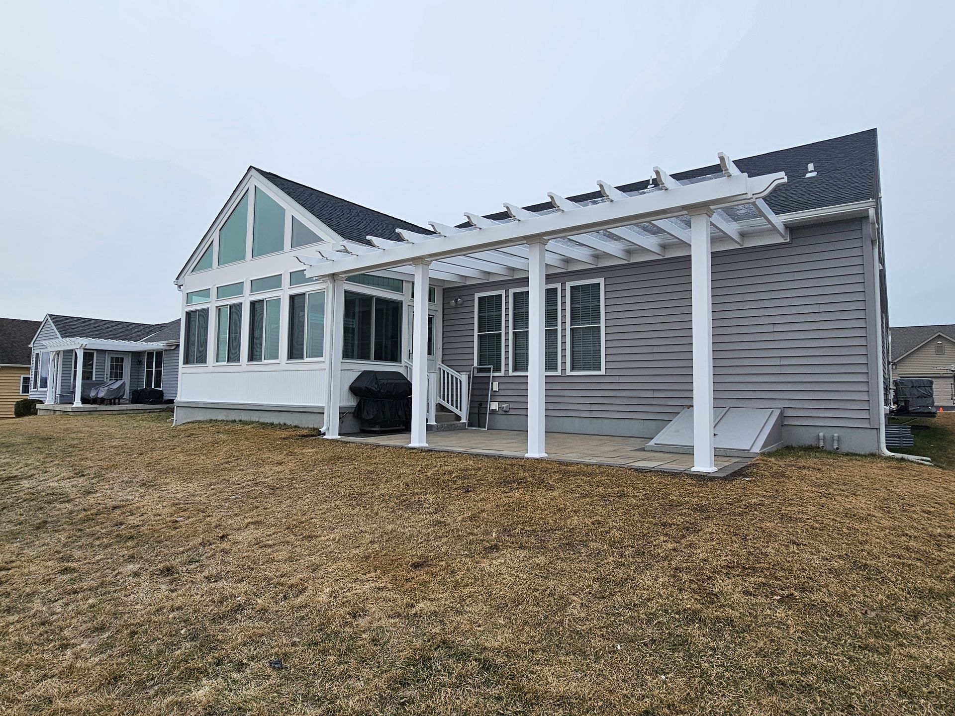 House with a white pergola, windows, and gray siding. Grassy yard under a cloudy sky.