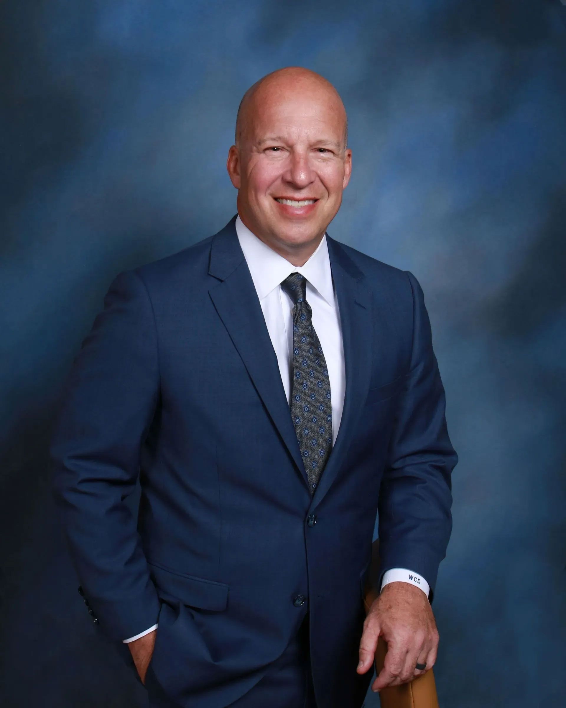 Smiling bald man in a navy suit with hands in pockets, posed against a blue studio backdrop.