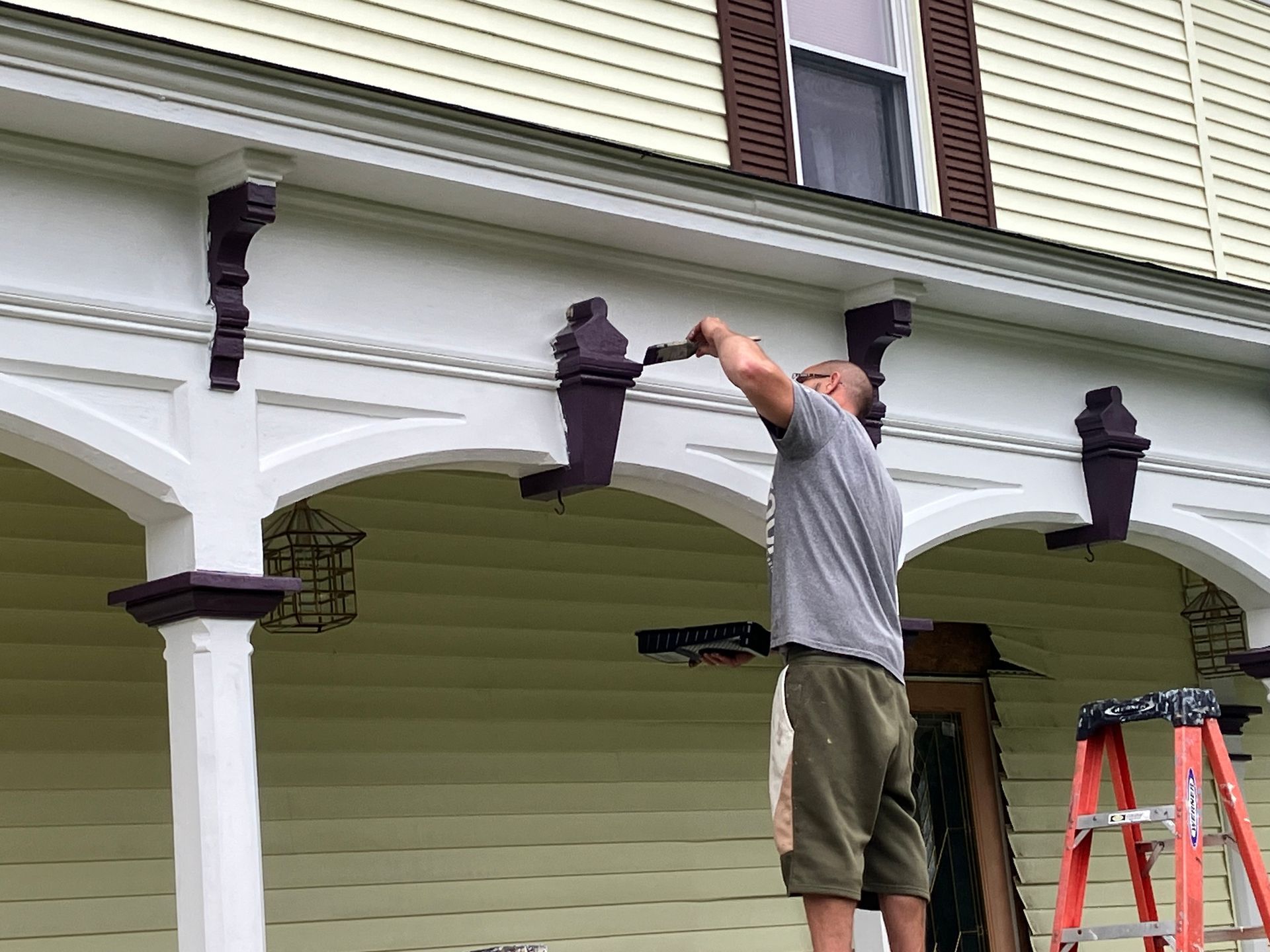 A man is standing on a ladder painting the porch of a house.