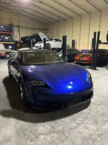 A blue Porsche Taycan parked in the foreground of a large garage featuring several other cars and storage lifts.